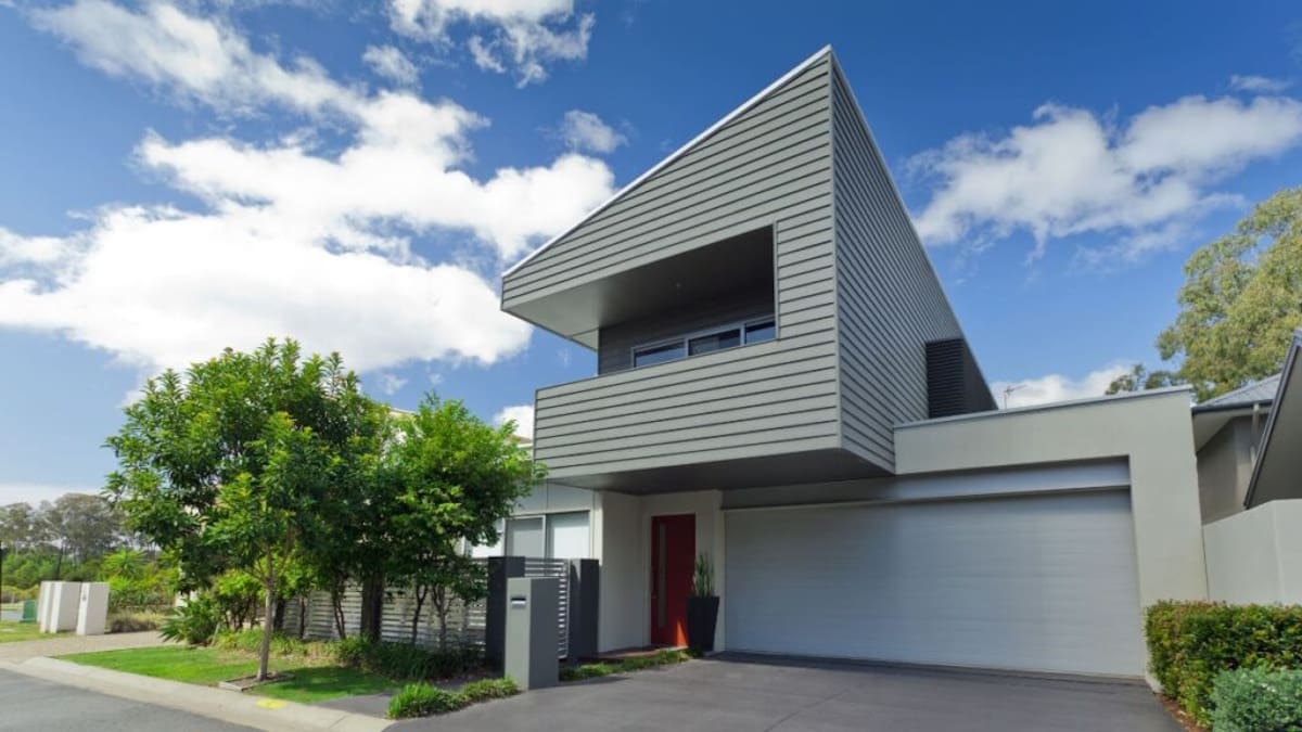 Een moderne woning met een uniek lessenaarsdak. Voor de woning zien we bomen en struiken, op de achtergrond de bewolkte lucht.