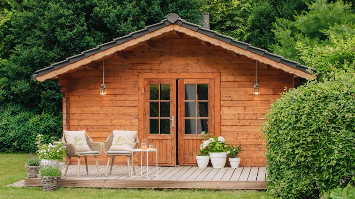 Een houten tuinhuis met dakpannen in een groene tuin. Voor het tuinhuis staan wat meubels.