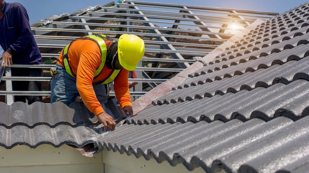 Professionele dakdekkers leggen grijze dakpannen op een dak. De dakdekker vooraan draagt een gele veiligheidshelm en beschermende kleding.