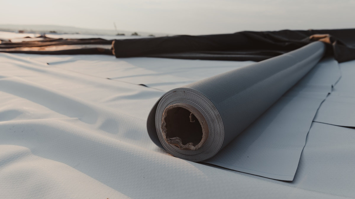 Een rol PVC dakbedekking ligt op een plat dak. De PVC is grijs aan de ene zijde en wit aan de andere zijde.